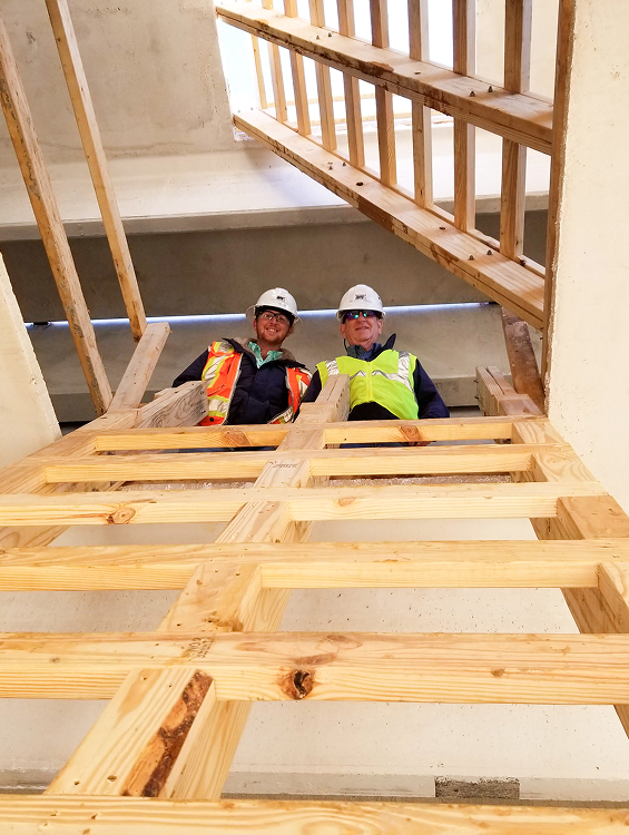 Two men wearing construction safety attire looking down an opening where wooden planks lean against the wall leading to the men
