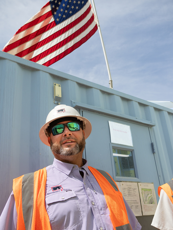 M&F employee wearing M&F branded hard hat and vest smiling in front of container box office with the American flag waving in the wind behind him